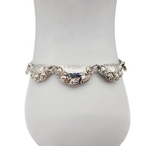 Könnte beinhalten: Ein silbernes Armband mit gehämmerter Oberfläche und einem floralen Design. Das Armband besteht aus einzelnen Gliedern, die die Form von Halbkreisen mit einem floralen Muster haben.