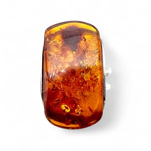 Puede incluir: Anillo rectangular de ámbar con una banda plateada. La piedra de ámbar es de un color marrón-anaranjado cálido y translúcido, con inclusiones visibles. El anillo está sobre un fondo blanco, resaltando los detalles de la joya.