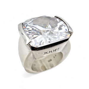Puede incluir: Un anillo plateado grande con una prominente piedra preciosa transparente de corte cuadrado. La banda del anillo es gruesa y tiene la palabra "JOOP!" grabada en el lateral. El anillo está sobre un fondo blanco.