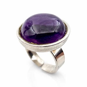 Könnte beinhalten: Ein silberfarbener Ring mit einem großen, glatten, ovalen violetten Edelstein in einer einfachen Fassung. Die Ringschiene ist an der Unterseite offen, was eine Anpassung ermöglicht, und das Metall hat eine polierte Oberfläche mit sichtbarer Textur.