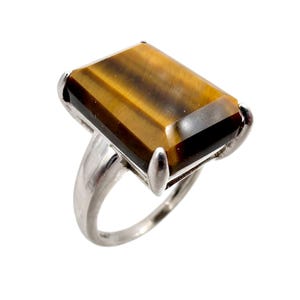 May include: A silver ring featuring a large, rectangular tiger's eye gemstone. The gemstone has a warm, brown color with golden streaks. The ring's band and setting are a bright silver color, creating a striking contrast with the stone.