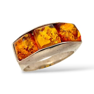 Könnte beinhalten: Ein Silberring mit drei rechteckigen Bernsteinsteinen. Die Bernsteine haben eine warme, durchscheinende orange Farbe und sind in einem gebogenen Silberband gefasst. Der Ring wird vor einem weißen Hintergrund präsentiert.