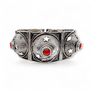 Könnte beinhalten: Ein silberfarbenes Armband mit drei großen, kreisförmigen, filigranverzierten Elementen. Jedes Element hat einen zentralen roten Stein und ist mit Sternen und geometrischen Formen verziert. Das Armband ist mit Scharnieren versehen.