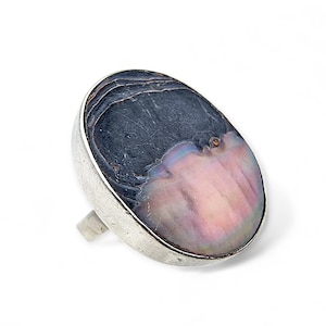 Könnte beinhalten: Ovaler Ring mit einem schillernden Stein in Grau-, Rosa- und Lilatönen, gefasst in einem silberfarbenen Metallband. Der Ring hat ein schlichtes, elegantes Design. Die Farben des Steins verändern sich im Licht.