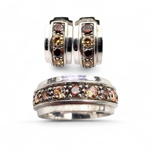 Könnte beinhalten: Ein silberner Ring und ein passendes Ohrring-Set. Der Ring und die Ohrringe haben eine Reihe kleiner, runder, mehrfarbiger Edelsteine in Braun-, Gelb- und Orangetönen. Der Schmuck ist vor einem weißen Hintergrund platziert.