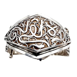 Könnte beinhalten: Ein silberfarbenes Armband mit einem verzierten, sich windenden Design. Das Armband hat ein zentrales Feld mit stilisierter Schrift und einer geometrischen Form. Das Metall hat ein leicht gealtertes Aussehen, und das Design ist aufwendig.