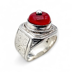 Könnte beinhalten: Silberner Ring mit einem roten, abgerundeten Stein. Der Ring hat ein verziertes, graviertes Design am Band und eine kleine silberne Kugel auf dem roten Stein. Der Ring ist vor einem weißen Hintergrund platziert.