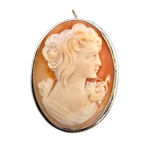 May include: Oval cameo pendant with a woman's profile carved in relief. The cameo is set in a silver-toned frame. The woman's portrait is in cream and peach tones, with detailed hair and floral accents.