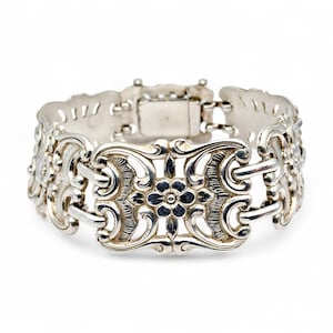 Könnte beinhalten: Ein silbernes Armband mit einem verzierten floralen Design. Das Armband hat eine Reihe von rechteckigen Gliedern mit aufwendigen Schnörkeln und einem zentralen Blumenmotiv. Der Verschluss ist eine einfache rechteckige Box. Das Armband wird vor weißem Hintergrund präsentiert.