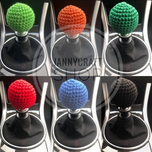May include: A collection of six crochet shift knobs in various colors: green, orange, green, red, blue, and black. Each knob is spherical and sits atop a black gear shift boot. The image includes the text "NANNYCRAFT SHOP".