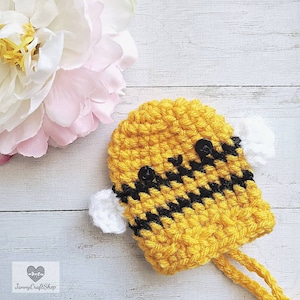 May include: A handmade crochet bee with yellow and black stripes, black button eyes, and white wings. The bee has two yellow strings attached. The background includes a white wooden surface and a pink and white flower.