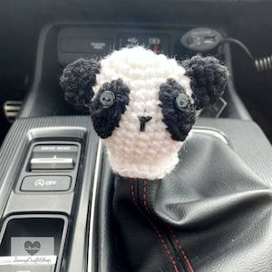 May include: A handmade, crocheted panda bear shift knob cover. The cover is white with black accents for the ears and eyes, and a small black nose. The shift knob cover is designed to fit over a car's gear shift.