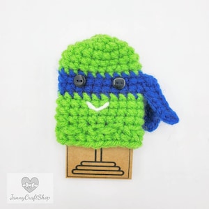 May include: A handmade crochet character in green and blue, featuring button eyes and a smiling mouth. The character has a blue headband and a matching tail. The item is displayed on a brown tag with a stand. The shop name "JannyCraftShop" is visible.