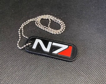 May include: A black metal dog tag with a white "N7" logo and a red triangle. The dog tag is attached to a silver chain.