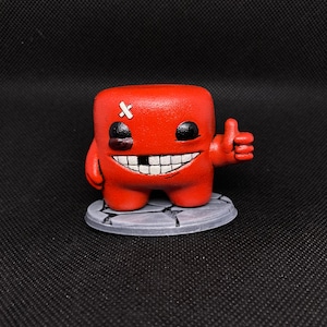 Könnte beinhalten: Rote, würfelförmige Figur mit einem breiten, zahnigen Grinsen und schwarzen Augen. Es hat einen Verband auf dem Kopf und zeigt einen Daumen hoch. Die Figur steht auf einem grauen, kreisförmigen Sockel.