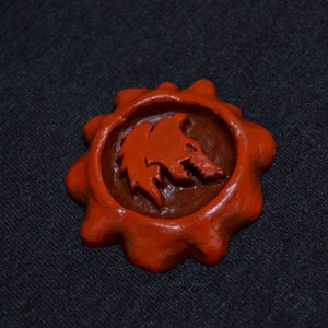 May include: A red wax seal with a detailed wolf head design. The seal is shaped like a starburst with a raised edge.