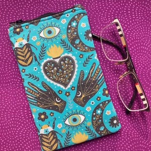 May include: A rectangular fabric pouch with a black zip closure featuring a teal background printed with black and gold illustrations of beetles, eyes, crescent moons, hearts, and hands, alongside a pair of spectacles with thin metal frames.