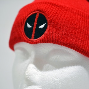 May include: Red knit beanie with a black and white Deadpool logo embroidered on the front.