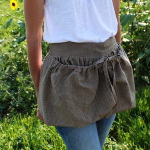 May include: A brown and white gingham apron with a gathered skirt and a tie at the waist. The apron is worn over a white t-shirt and blue jeans.