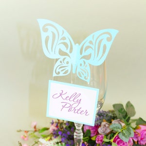May include: A light blue paper butterfly place card holder with a white card that says "Kelly Porter" in purple script. The place card holder is on a wine glass with a bouquet of purple and pink flowers.