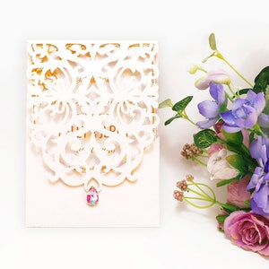 May include: White laser cut wedding invitation with a pink and gold design. A single rhinestone embellishment is attached to the center of the invitation.