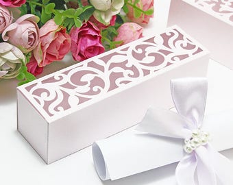 May include: White cardboard box with a delicate, intricate, pink floral design cut out. The box is open and a white scroll with a white satin ribbon and pearl embellishments is inside.
