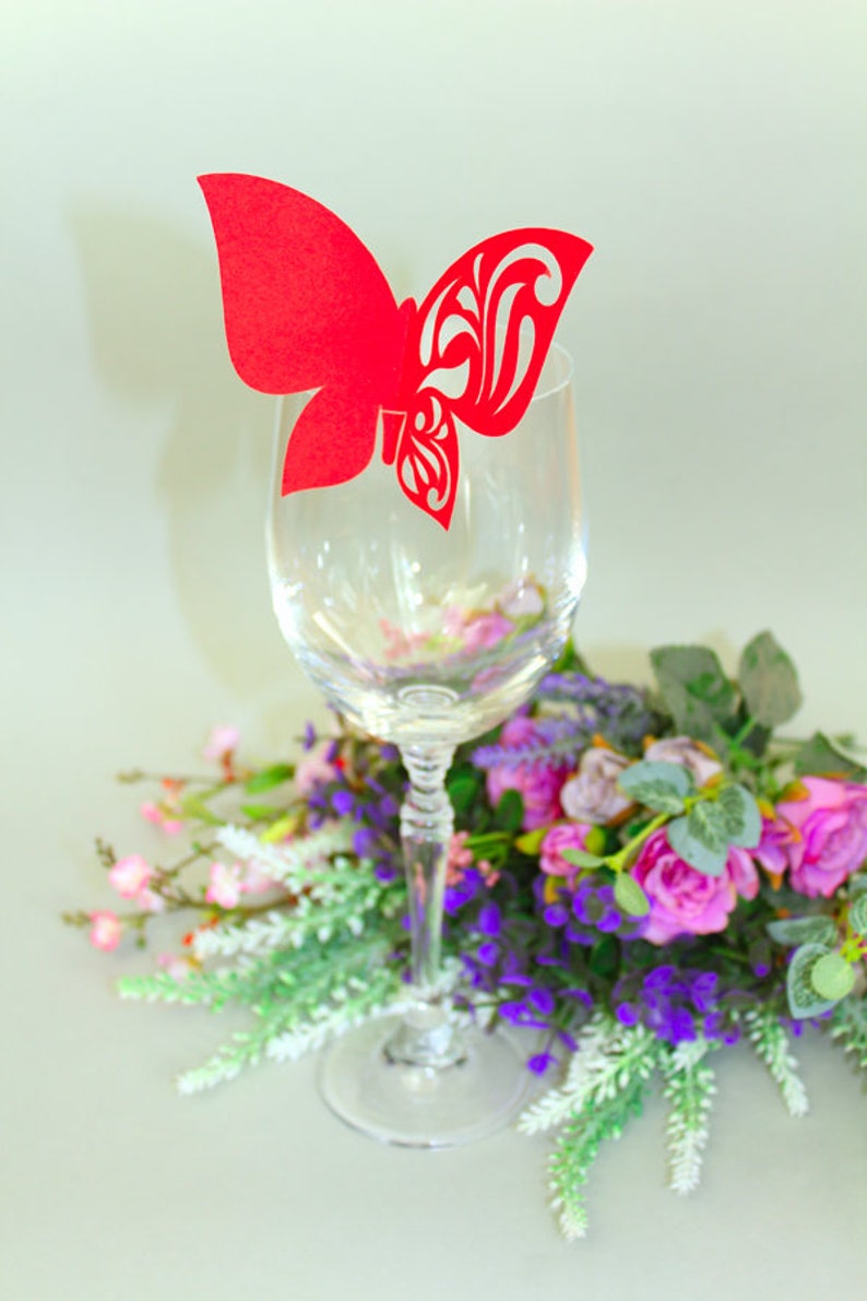 May include: A red paper butterfly place card holder on a clear glass wine glass. The glass is surrounded by a bouquet of pink and purple flowers.