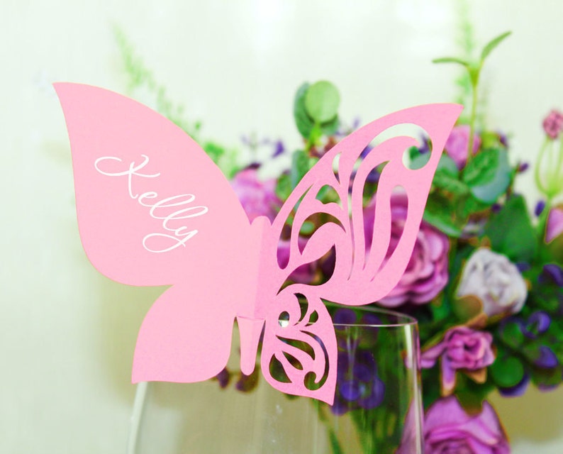 May include: Pink paper butterfly place card with the name "Kelly" written in white calligraphy. The butterfly is attached to a clear glass stem.