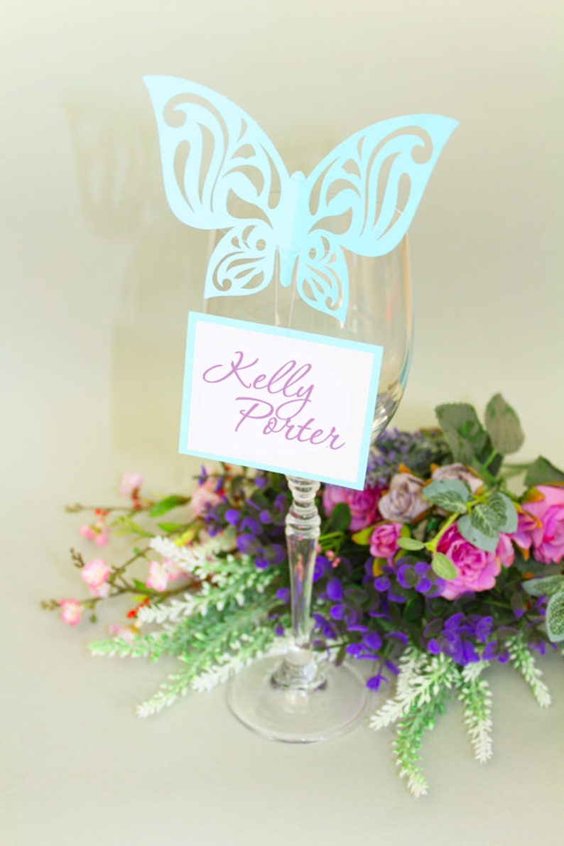 May include: A light blue paper butterfly place card holder with a white card that says "Kelly Porter" in purple script. The place card holder is on a wine glass with a bouquet of purple and pink flowers.