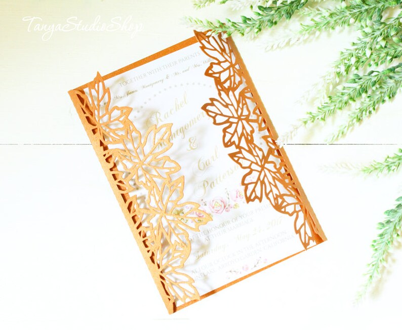 Puede incluir: Invitaci&oacute;n de boda de corte l&aacute;ser dorado con un dise&ntilde;o de hojas. La invitaci&oacute;n est&aacute; doblada por la mitad y las hojas est&aacute;n recortadas en el panel frontal. La invitaci&oacute;n est&aacute; sobre un fondo blanco.