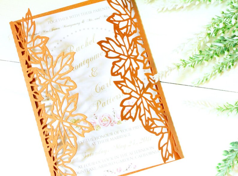 Puede incluir: Una invitaci&oacute;n de boda cortada con l&aacute;ser con un tema oto&ntilde;al. La invitaci&oacute;n es naranja con fondo blanco y presenta un dise&ntilde;o de hojas alrededor de los bordes. El texto de la invitaci&oacute;n dice "Rachel & Carl".