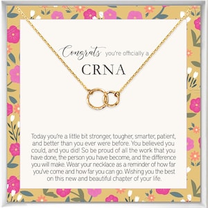 May include: A gold necklace with two interlocking circles, presented in a gift box with a floral pattern. The box includes a congratulatory message for a new CRNA, saying "Congrats you're officially a CRNA". The message continues with words of encouragement and pride for the recipient's accomplishments.