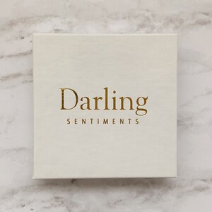 Puede incluir: Una caja blanca con el texto dorado "Darling Sentiments" en la parte delantera.