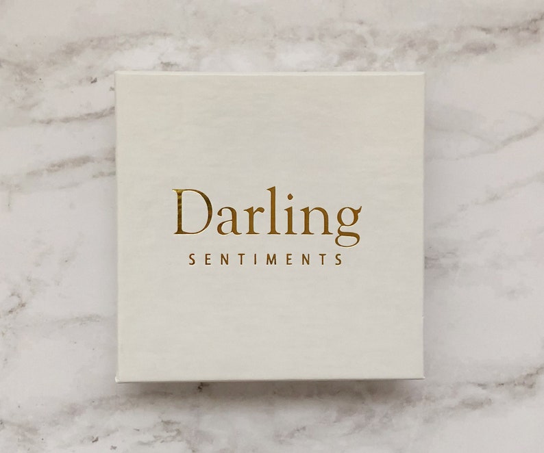 Puede incluir: Una caja blanca con el texto dorado "Darling Sentiments" en la parte delantera.