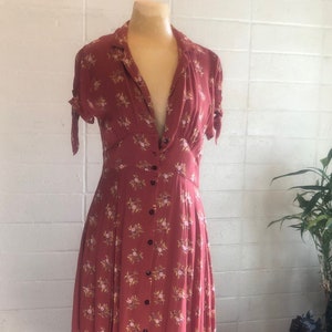 Peut inclure: Une robe maxi de couleur rouille &agrave; imprim&eacute; floral et &agrave; fermeture boutonn&eacute;e sur le devant. La robe a un col en V et des manches courtes avec des liens aux poignets. L'ourlet est asym&eacute;trique, avec un dos plus long et un devant plus court.