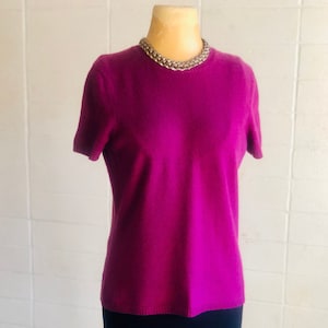 May include: A magenta short-sleeve sweater and a black skirt are displayed on a mannequin. A gold-colored chain necklace is around the mannequin's neck. The items are on a wooden stand against a white tiled wall.