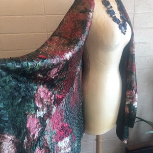 May include: A floral patterned shawl in shades of green, red, and pink is draped over a mannequin. The fabric has a textured, quilted appearance. A blue necklace is around the mannequin's neck. The shawl is displayed in a well-lit room.