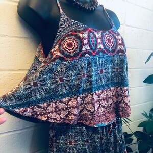 Peut inclure: Une combinaison sans manches avec un corsage superposé et un motif multicolore composé de formes géométriques et florales dans des tons bleu sarcelle, bordeaux et blanc. Le vêtement possède de fines bretelles et une coupe décontractée.
