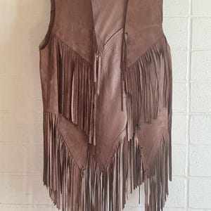 May include: Brown suede vest with long fringe detailing. The vest hangs on a wooden hanger against a white brick wall. The vest has a V-neck and a flowing, open front design.