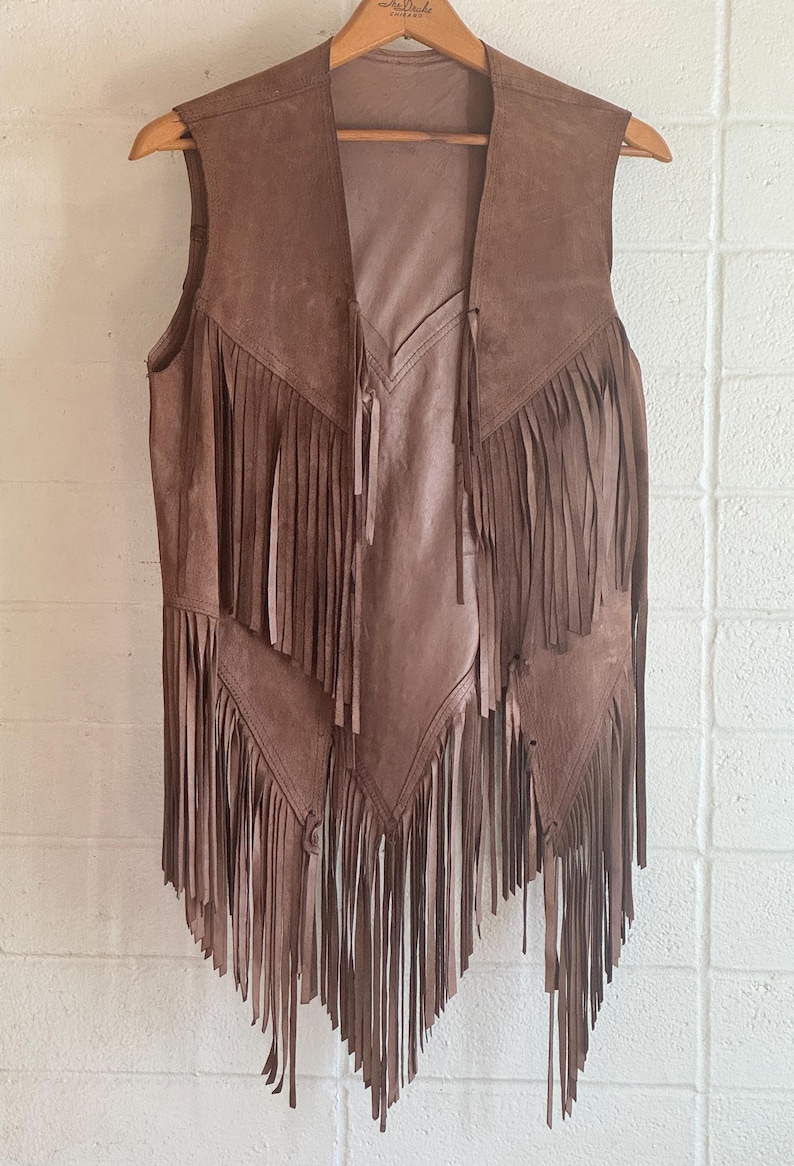 May include: Brown suede vest with long fringe detailing. The vest hangs on a wooden hanger against a white brick wall. The vest has a V-neck and a flowing, open front design.