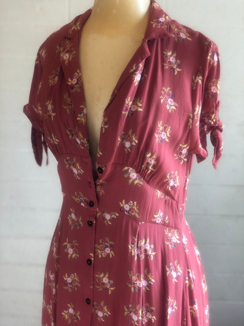 Peut inclure: Une longue robe &agrave; fleurs rouge &agrave; imprim&eacute; floral avec un devant boutonn&eacute; et des manches courtes. La robe a un corsage ajust&eacute; et une jupe ample.