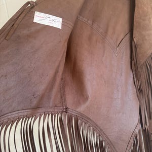 May include: Brown leather vest with fringe detailing. The vest has a V-neck design and is hanging on a wooden hanger. The interior label reads "BOUTIQUE Y.O. PARIS".