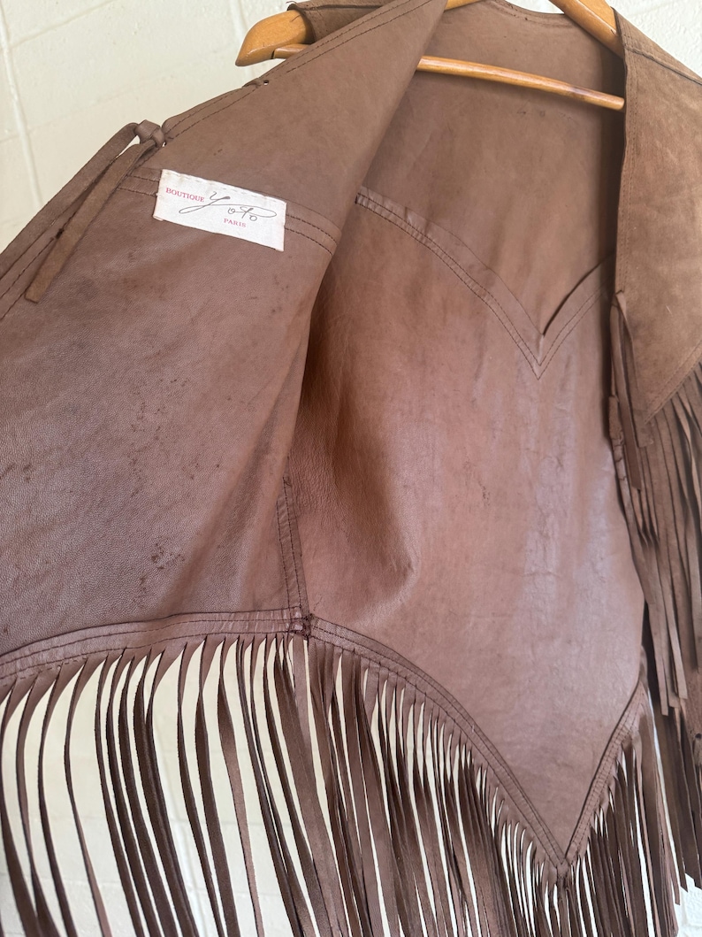 May include: Brown leather vest with fringe detailing. The vest has a V-neck design and is hanging on a wooden hanger. The interior label reads "BOUTIQUE Y.O. PARIS".