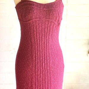 May include: A burgundy-coloured, ribbed knit dress with a fitted bodice and spaghetti straps. The dress has a form-fitting silhouette, showcasing a vertical ribbed texture throughout. The dress is displayed on a mannequin.