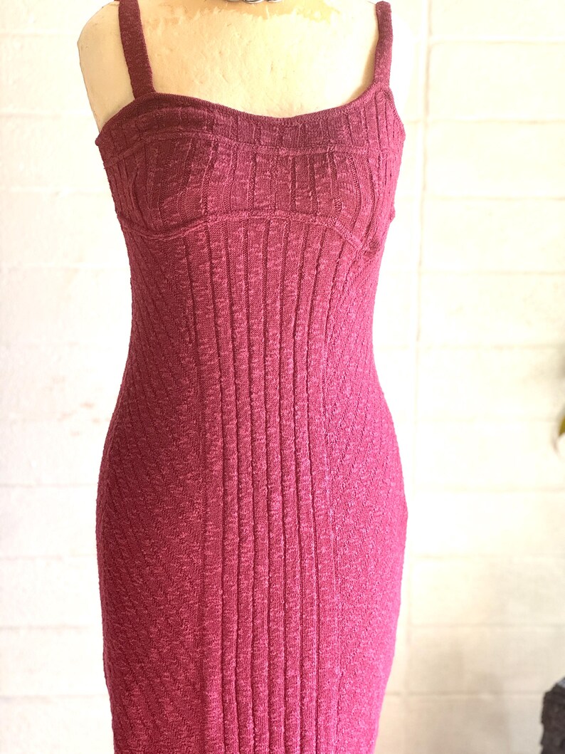 May include: A burgundy-coloured, ribbed knit dress with a fitted bodice and spaghetti straps. The dress has a form-fitting silhouette, showcasing a vertical ribbed texture throughout. The dress is displayed on a mannequin.