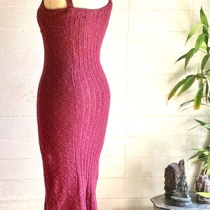 May include: A burgundy-coloured, ribbed knit dress on a mannequin. The dress has a square neckline, thin straps, and a fitted shape that flares at the hem. A silver chain necklace is around the mannequin's neck.