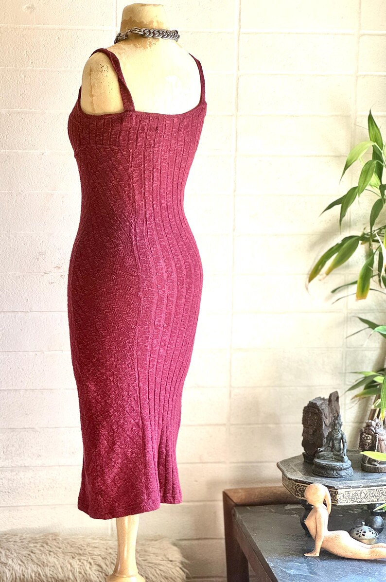 May include: A burgundy-coloured, ribbed knit dress on a mannequin. The dress has a square neckline, thin straps, and a fitted shape that flares at the hem. A silver chain necklace is around the mannequin's neck.