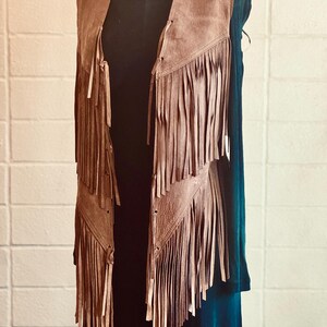 May include: A brown suede vest with long fringe detailing. The vest is displayed on a mannequin, showcasing its open front and layered fringe design. A silver chain necklace is visible at the neckline. The vest is paired with a black top and a teal dress.