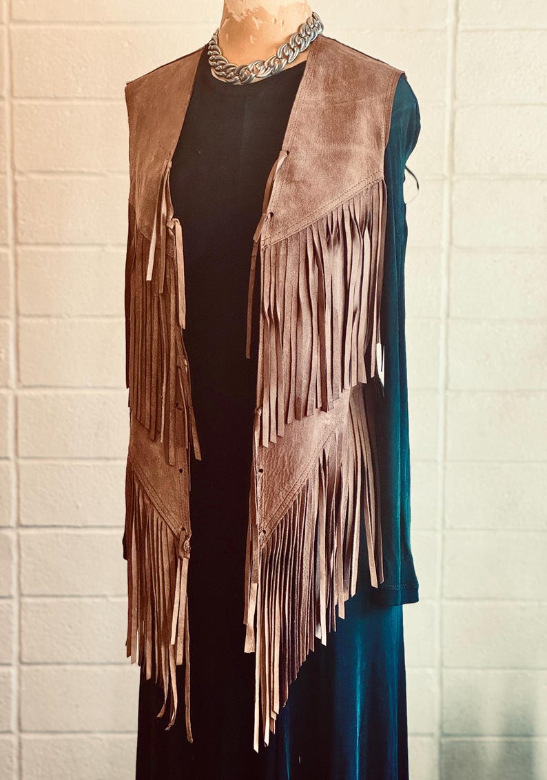 May include: A brown suede vest with long fringe detailing. The vest is displayed on a mannequin, showcasing its open front and layered fringe design. A silver chain necklace is visible at the neckline. The vest is paired with a black top and a teal dress.