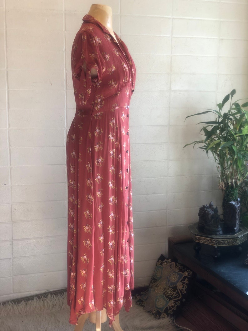 Peut inclure: Une longue robe de couleur rouille &agrave; imprim&eacute; floral. La robe a un devant boutonn&eacute;, un col en V et des manches courtes. La robe est sur un mannequin.